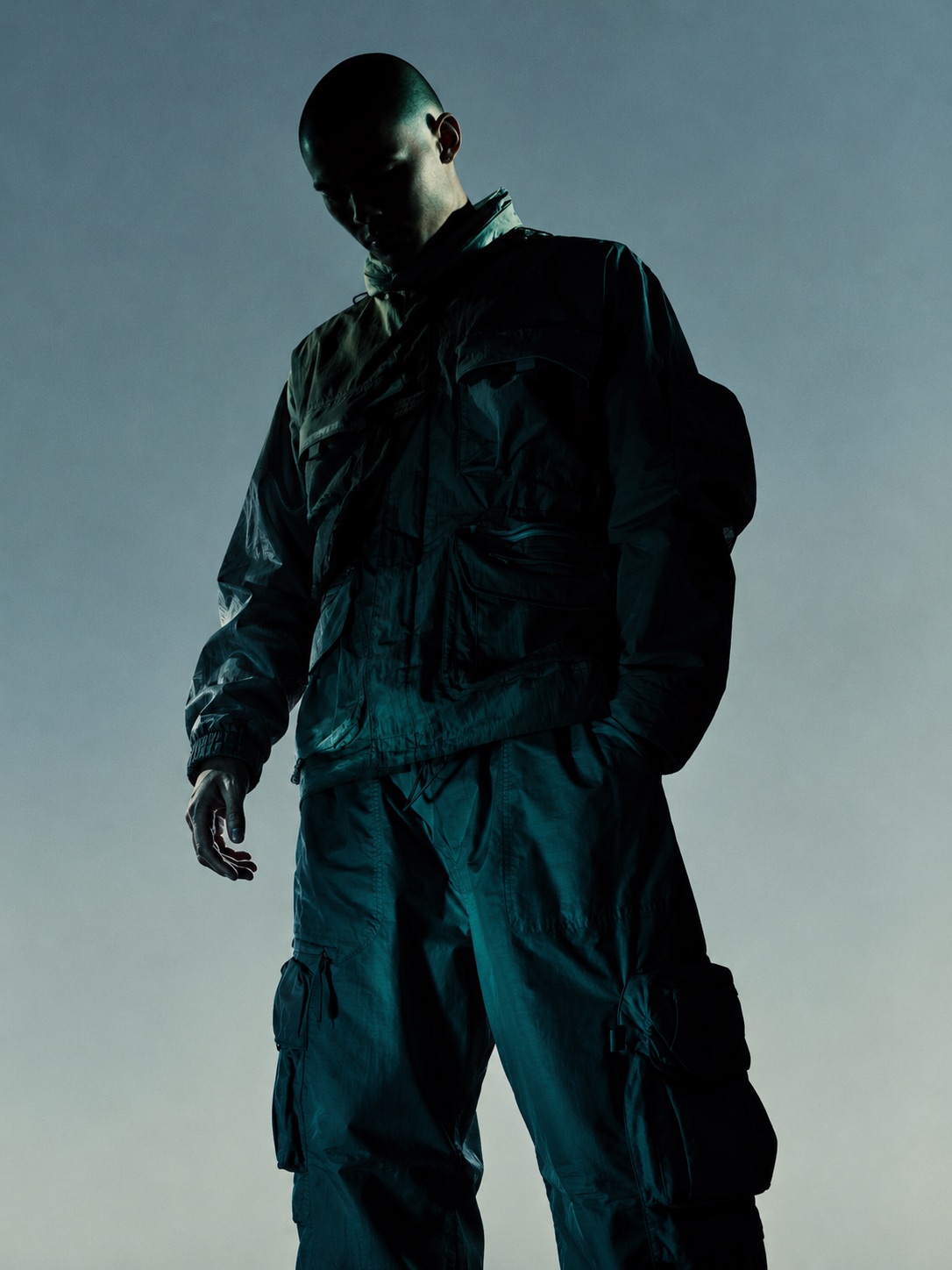 Figure in black technical streetwear looking down at a phone, backlit silhouette