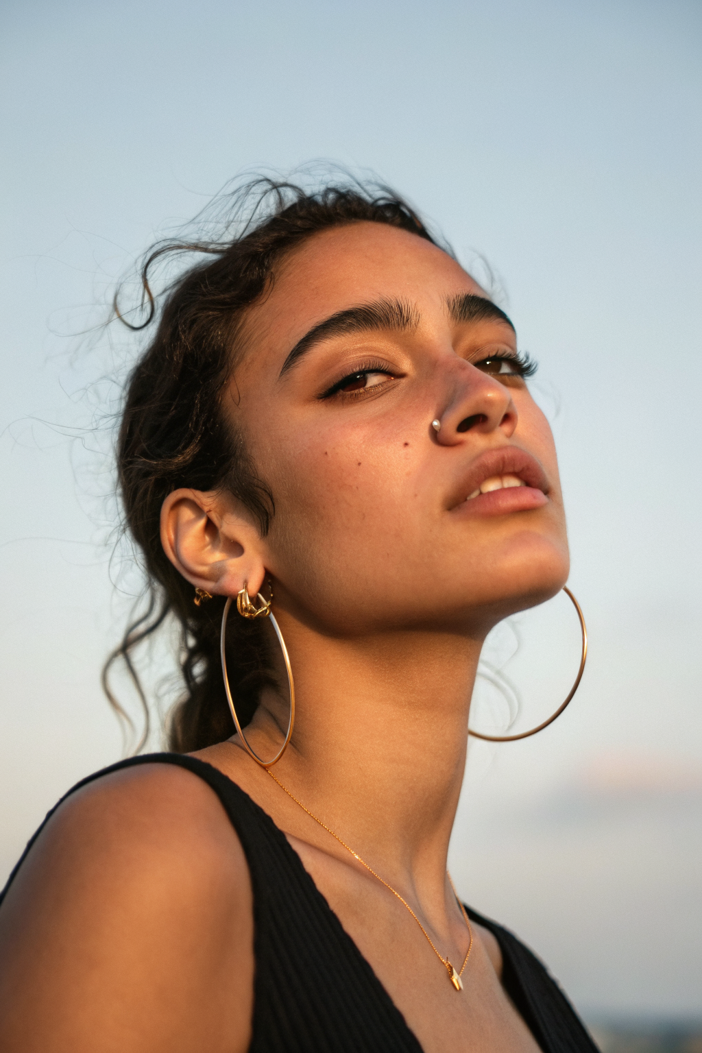 Editorial portrait of a woman in silhouette with gold hoop earrings against a pale sky, holding a small glowing UI element
